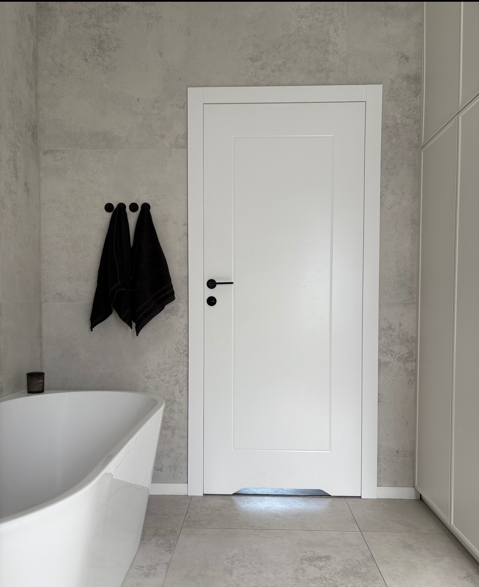 Białe, minimalistyczne drzwi wewnętrzne z czarną klamką w łazience z szarymi płytkami i wanną. Dwa czarne ręczniki wiszą na wieszaku.