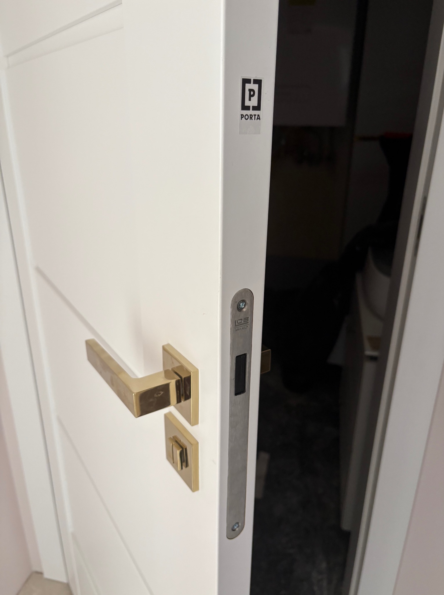 Białe drzwi wewnętrzne Porta z złotą klamką i srebrnym zamkiem, widok z bliska na detal wykończenia i otwarte skrzydło.