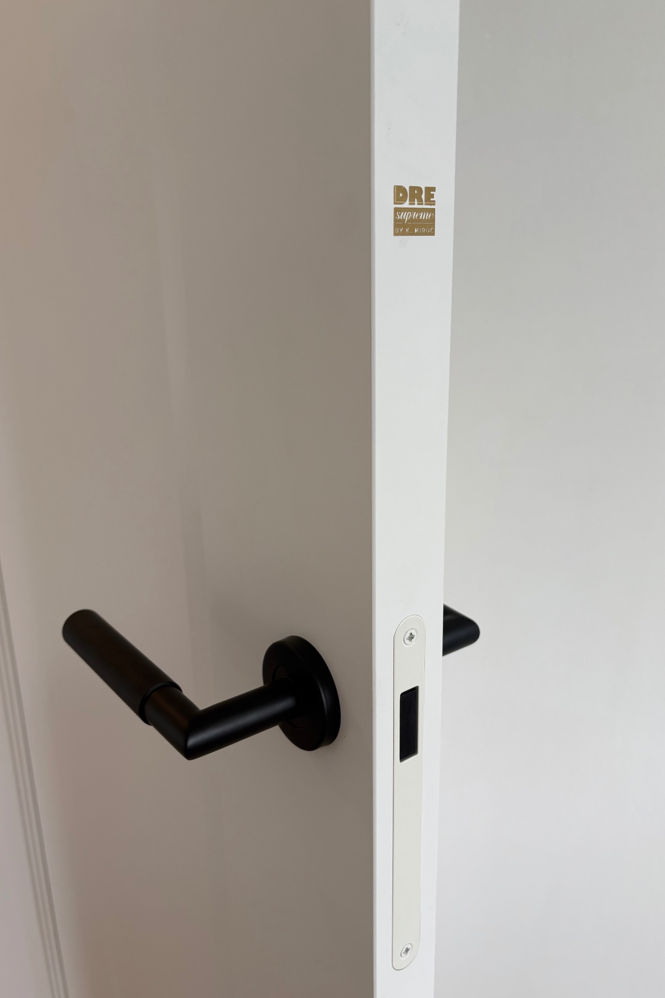Białe drzwi wewnętrzne z czarną klamką i zamkiem wpuszczanym, widoczne logo DRE Supreme, minimalistyczny design.