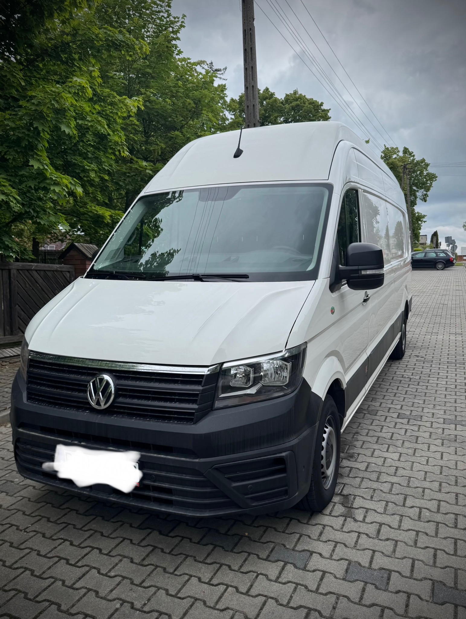 Biały Volkswagen Crafter z wysokim dachem, zaparkowany na brukowanej ulicy, w tle drzewa i słup elektryczny. Auto czeka na załadunek przed przeprowadzką.