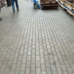 SVRUSM SPÓŁKA Z OGRANICZONĄ ODPOWIEDZIALNOŚCIĄ - Ułożona kostka brukowa na posesji z pracownikami w tle. Widoczny wózek z paletami i budynek mieszkalny. Świeżo ułożona, mokra kostka.