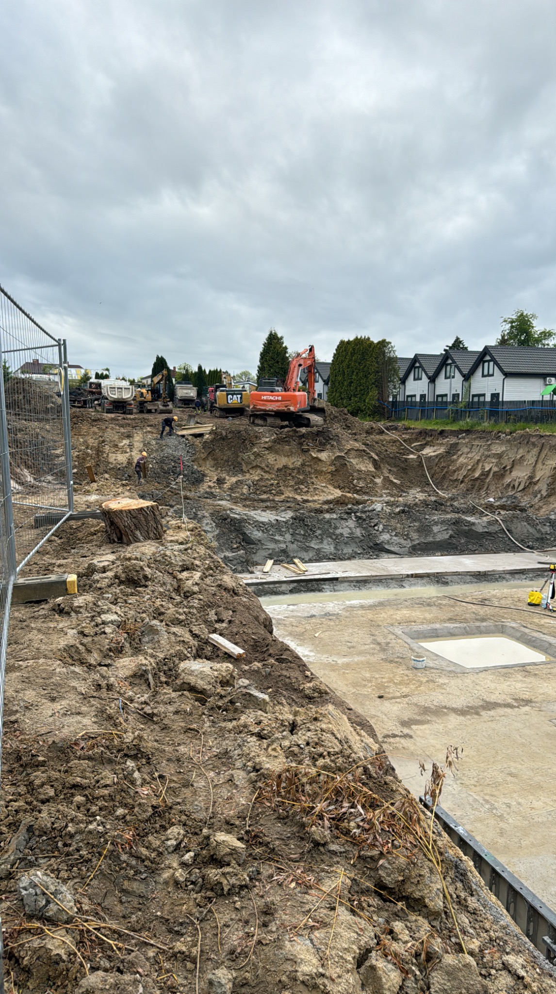 Rozległy wykop z pracującymi koparkami CAT i Hitachi na tle zabudowy mieszkalnej pod pochmurnym niebem. Widoczny fragment ogrodzenia i wykopane fundamenty.