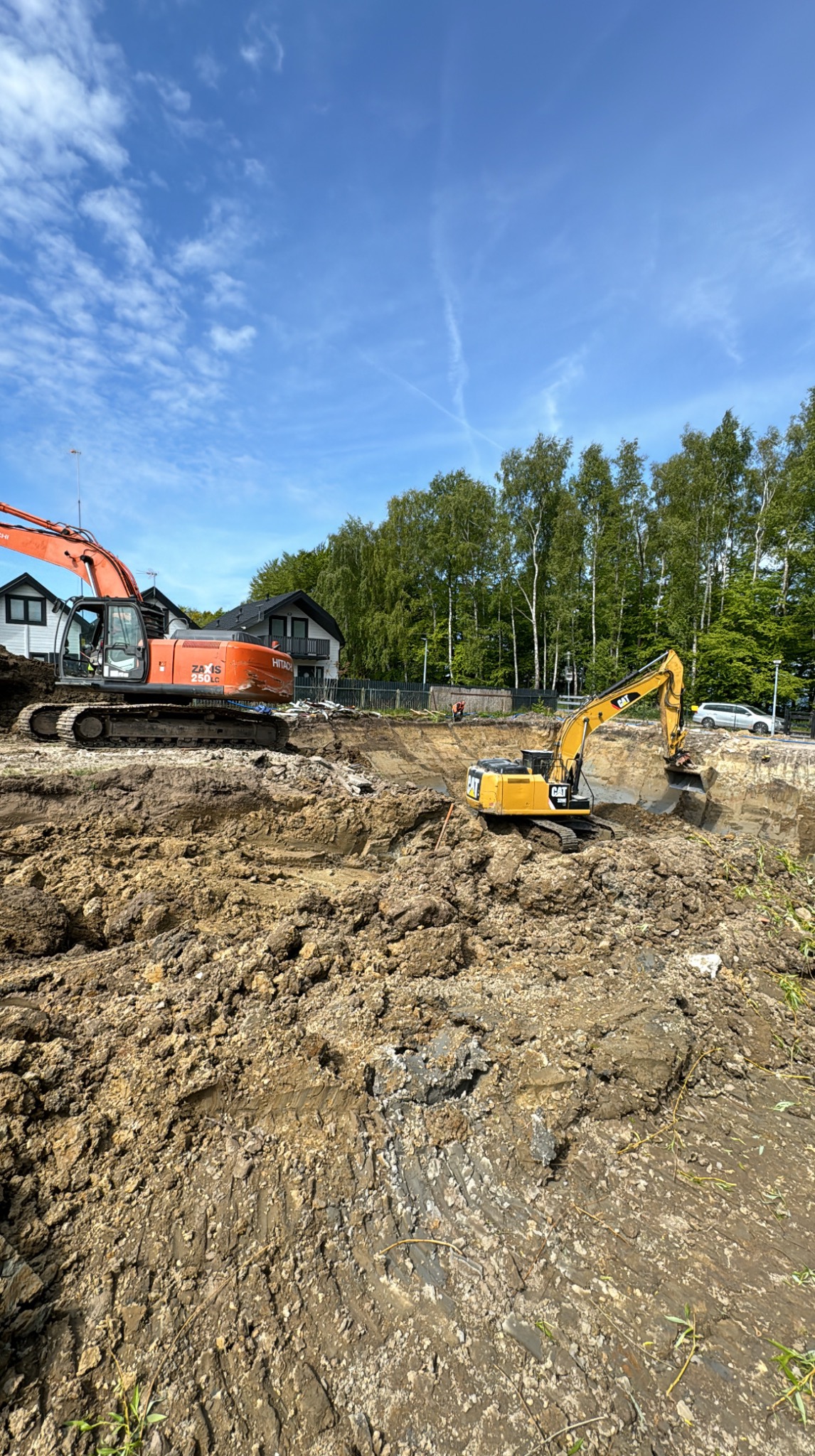 Dwie koparki (pomarańczowa Hitachi i żółta CAT) pracują w głębokim wykopie na tle domów i drzew. Błękitne niebo z delikatnymi chmurami w tle.