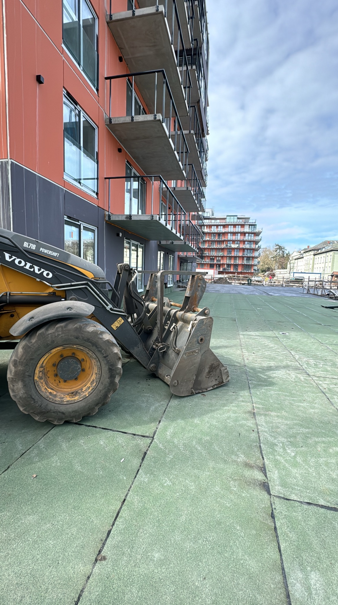 Żółta ładowarka Volvo BL71B na zielonej powierzchni, w tle budynek z balkonami i kolejny budynek w oddali, plac budowy, dzień.