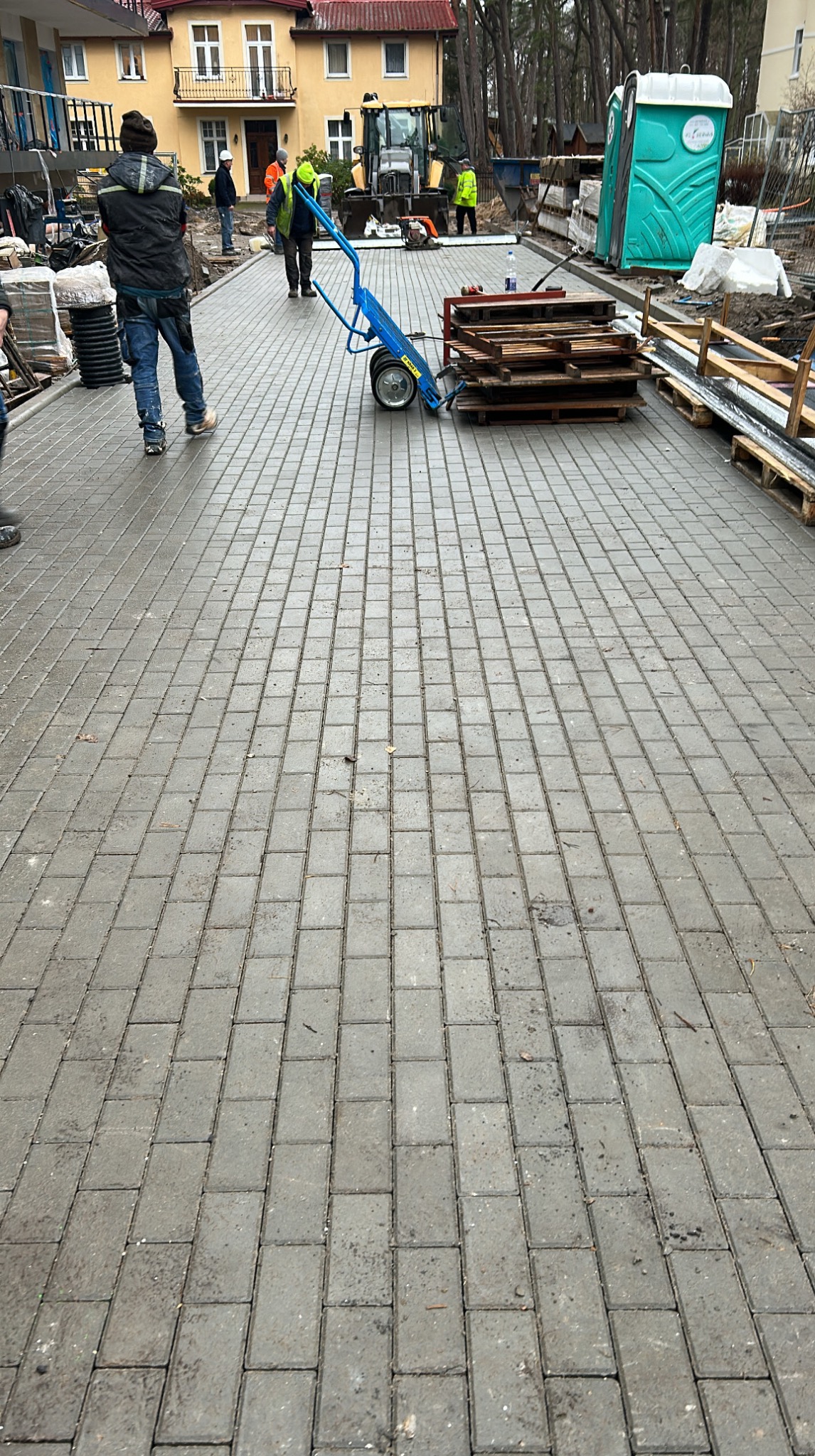 Ułożona kostka brukowa na posesji z pracownikami w tle. Widoczny wózek z paletami i budynek mieszkalny. Świeżo ułożona, mokra kostka.