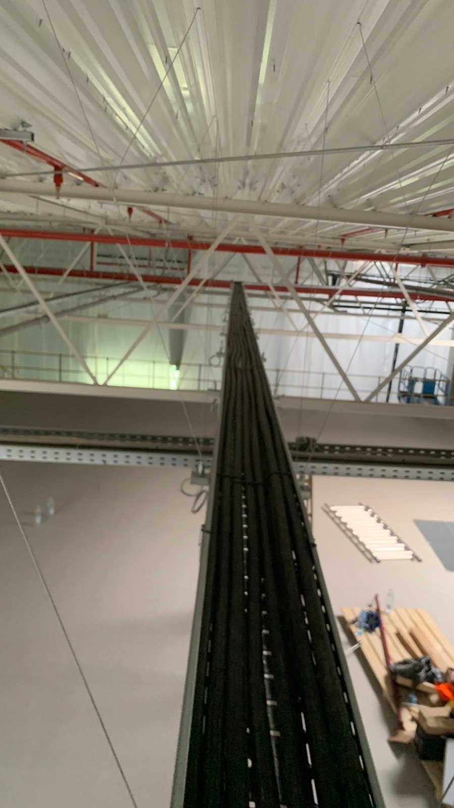 Wysoka kolumna kabli w czarnych osłonach, biegnąca od podłogi hali do sufitu z instalacją oświetleniową i przeciwpożarową. Widoczne rusztowania i paleta.