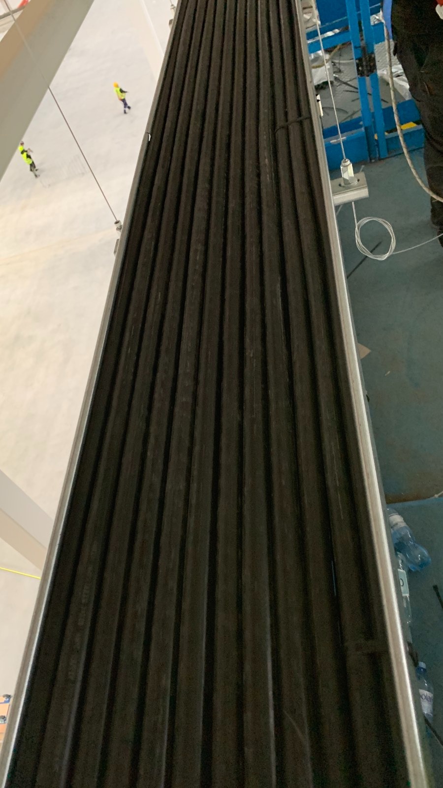 Czarna, żebrowana rura osłonowa, prawdopodobnie element instalacji odgromowej, zamocowana na metalowej konstrukcji w hali przemysłowej. Widoczne liny mocujące.