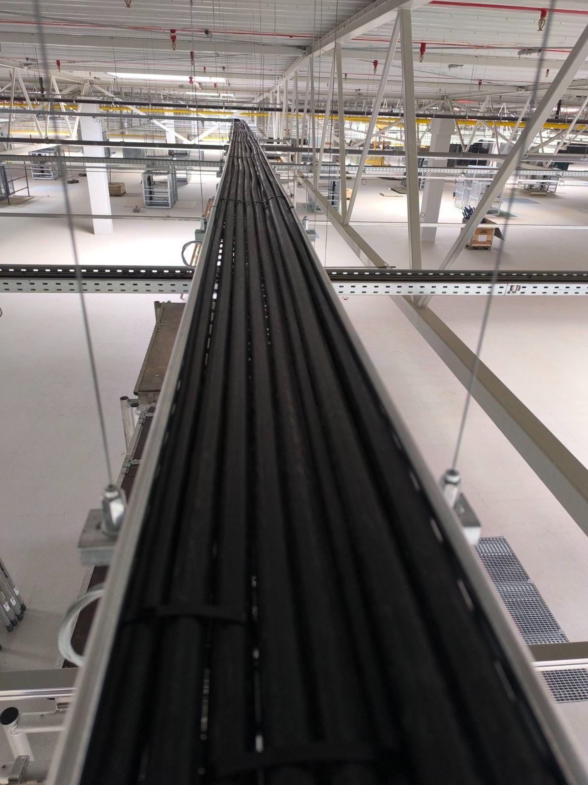Równolegle ułożone czarne kable w korycie na hali produkcyjnej, widok z góry, oświetlenie sufitowe, metalowa konstrukcja wsporcza.