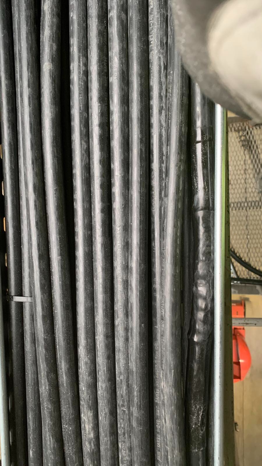 Rząd czarnych kabli elektrycznych w metalowej ramie, jeden kabel z widocznym łączeniem izolacyjnym. Ujęcie z bliska, detale instalacji.