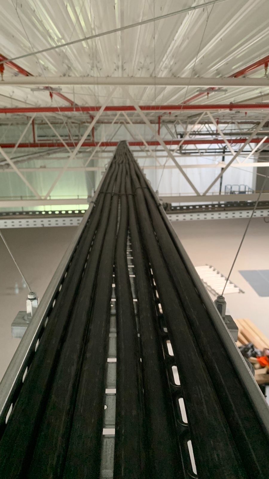 Wiązka kabli elektrycznych prowadzona w korytach metalowych pod sufitem hali przemysłowej. Widoczne elementy konstrukcji dachu i oświetlenie.