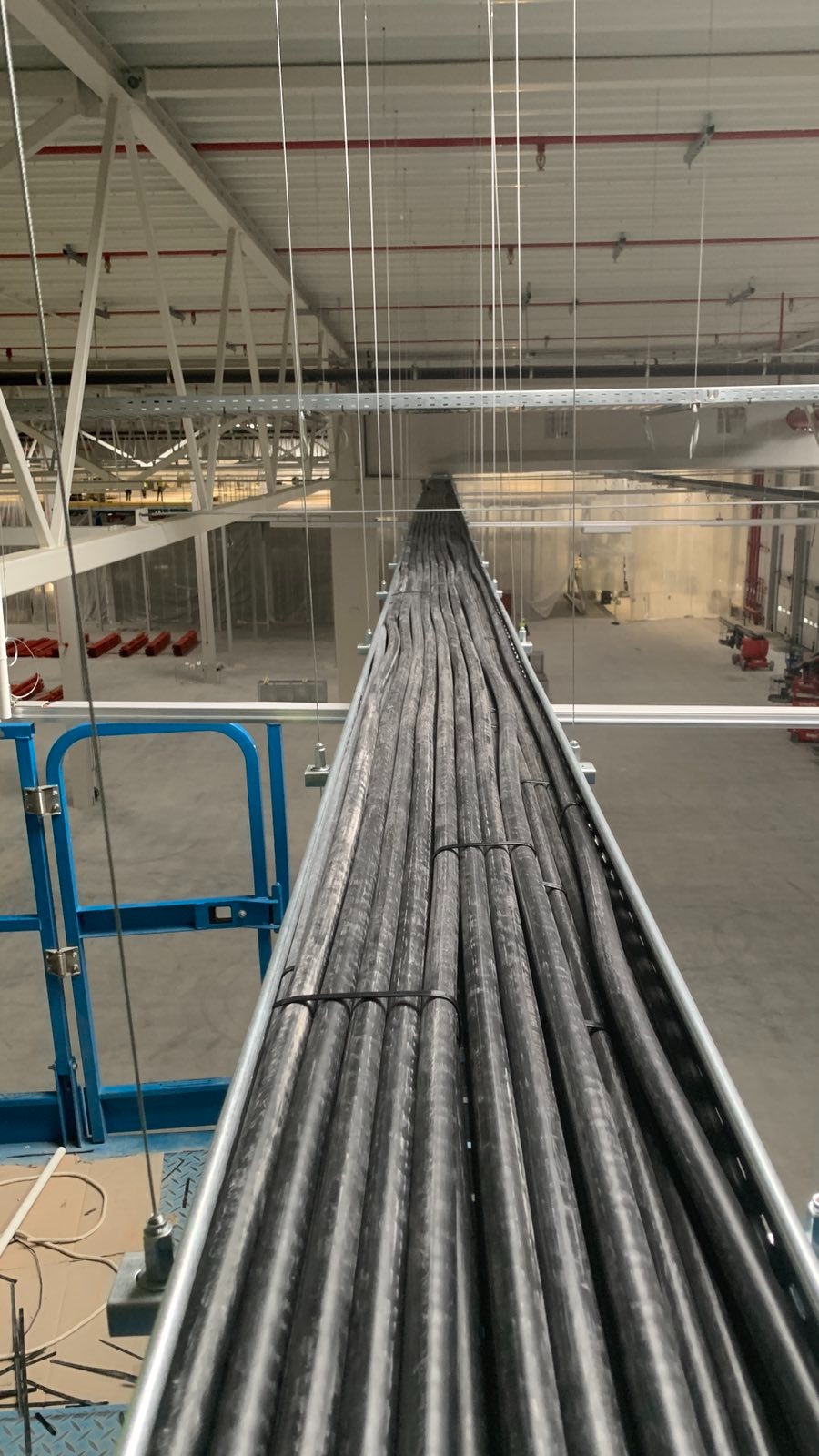 Rząd kabli elektrycznych ułożonych w metalowej korycie, zawieszonych pod sufitem hali przemysłowej. Widok z podnośnika koszowego na instalację.