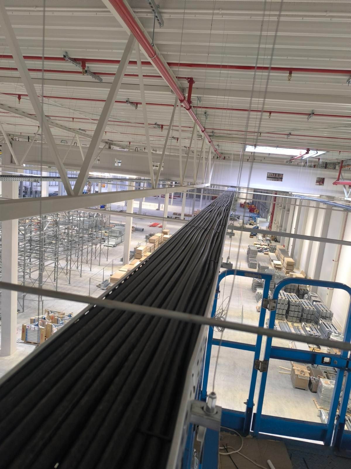 Kable elektryczne ułożone w korycie na wysokości w hali magazynowej. Widok z podnośnika koszowego, instalacja w trakcie realizacji, widoczne elementy konstrukcji hali.