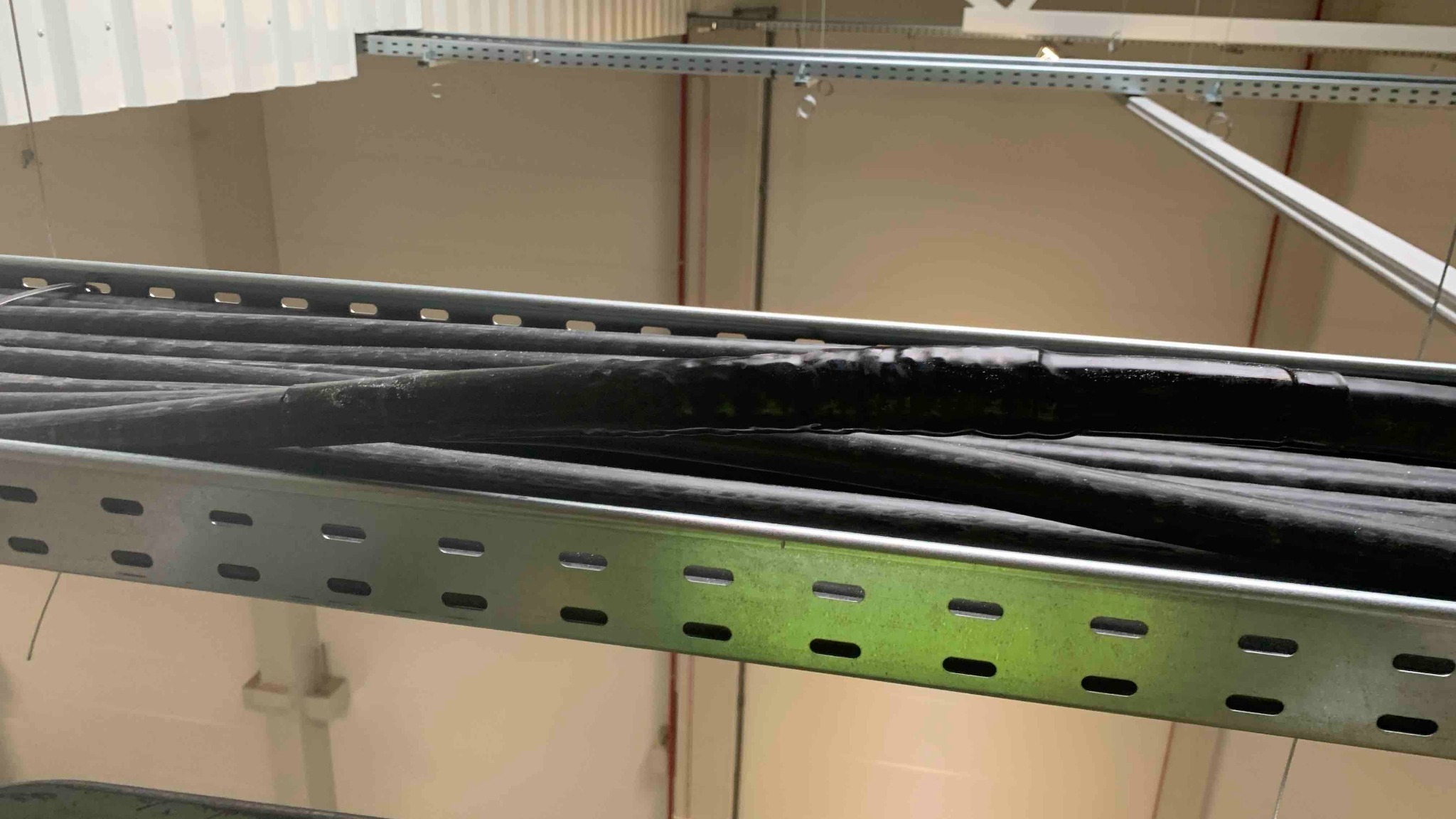 Ułożone kable elektryczne w metalowej korycie pod sufitem, widoczne łączenia zabezpieczone taśmą izolacyjną, oświetlenie przemysłowe w tle.