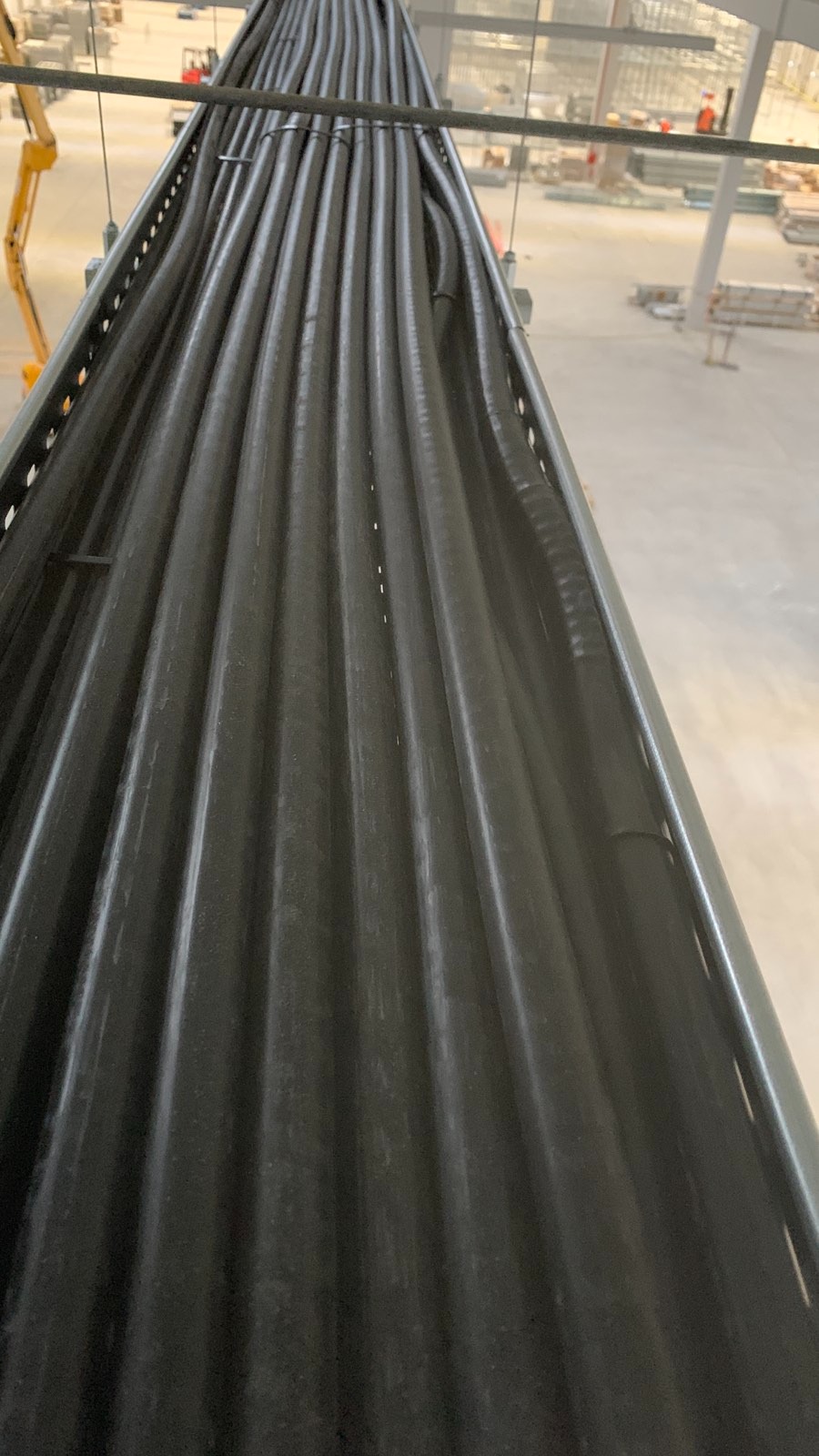 Rząd czarnych, karbowanych rur prowadzonych w metalowej korycie, widok z góry, w tle plac budowy z maszynami i pracownikami.