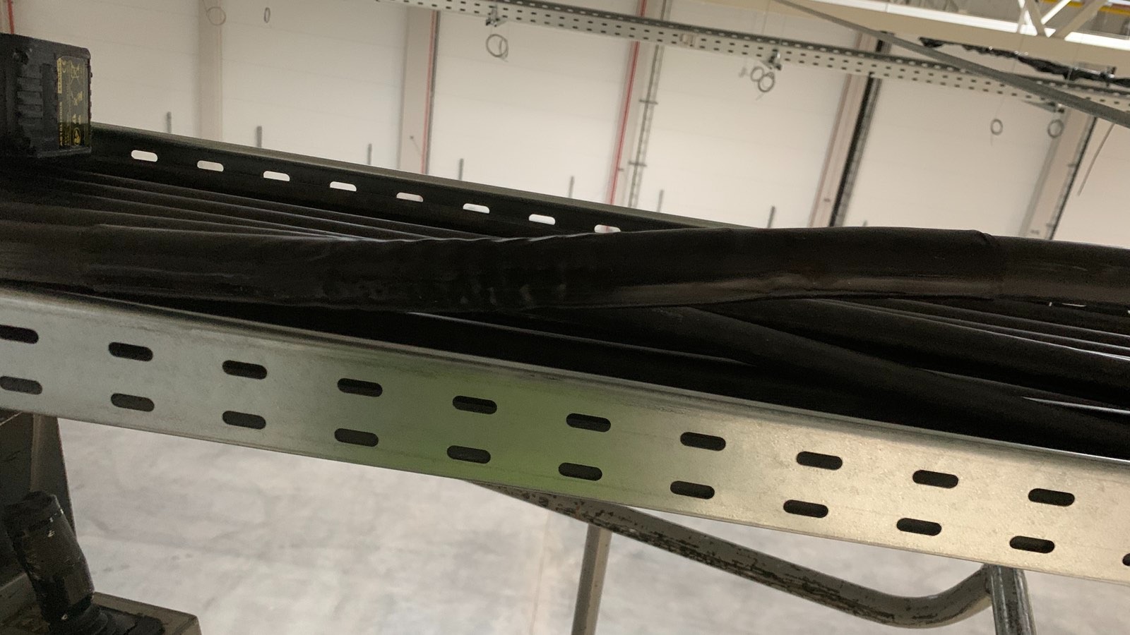 Ułożone kable elektryczne w korycie kablowym, widok z dołu w hali magazynowej w Olsztynie. Szare koryto z perforacją, czarne przewody.