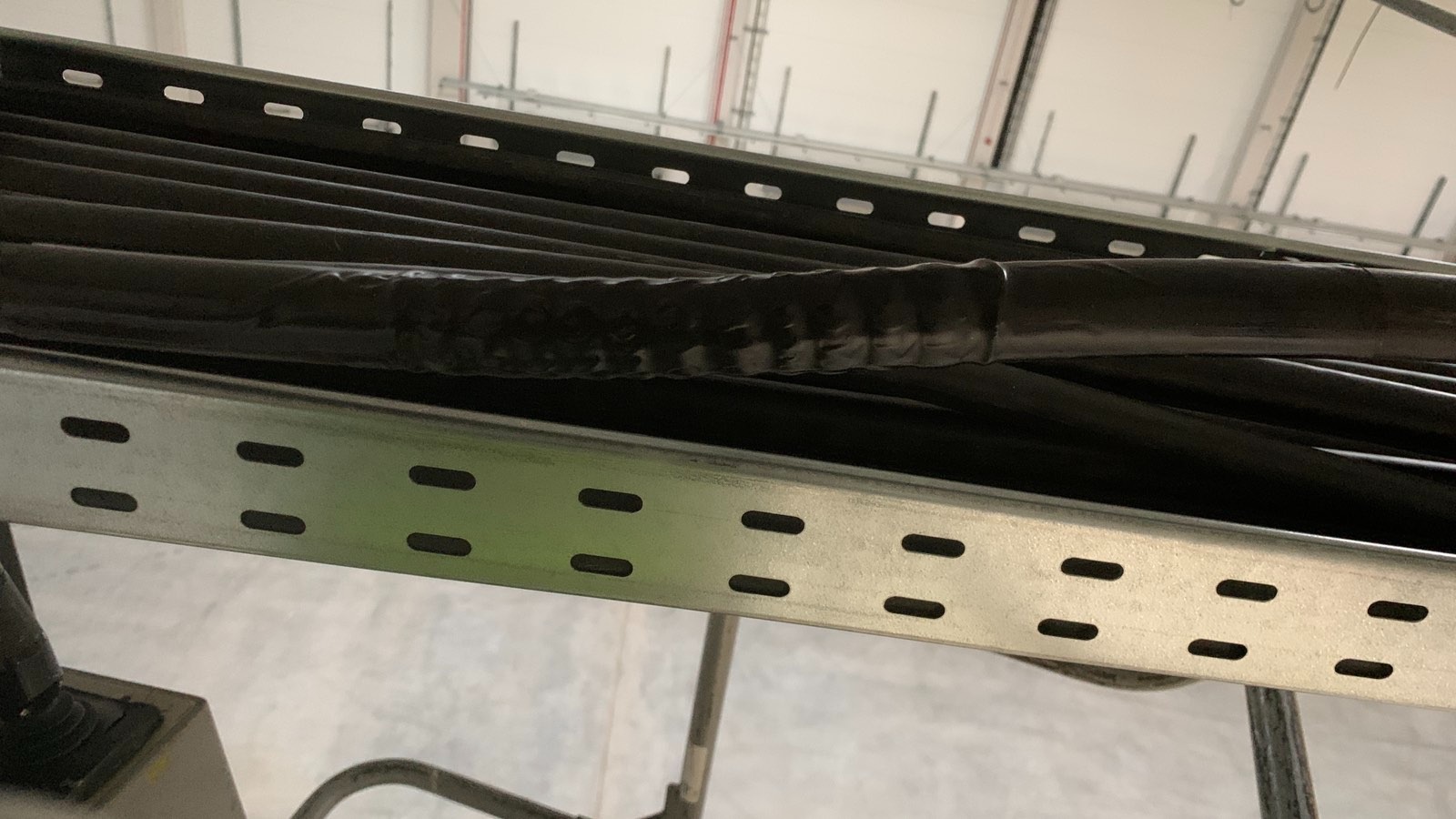 Ułożone kable elektryczne w metalowej korycie, zabezpieczone taśmą izolacyjną, widoczne elementy mocowania do sufitu w tle.