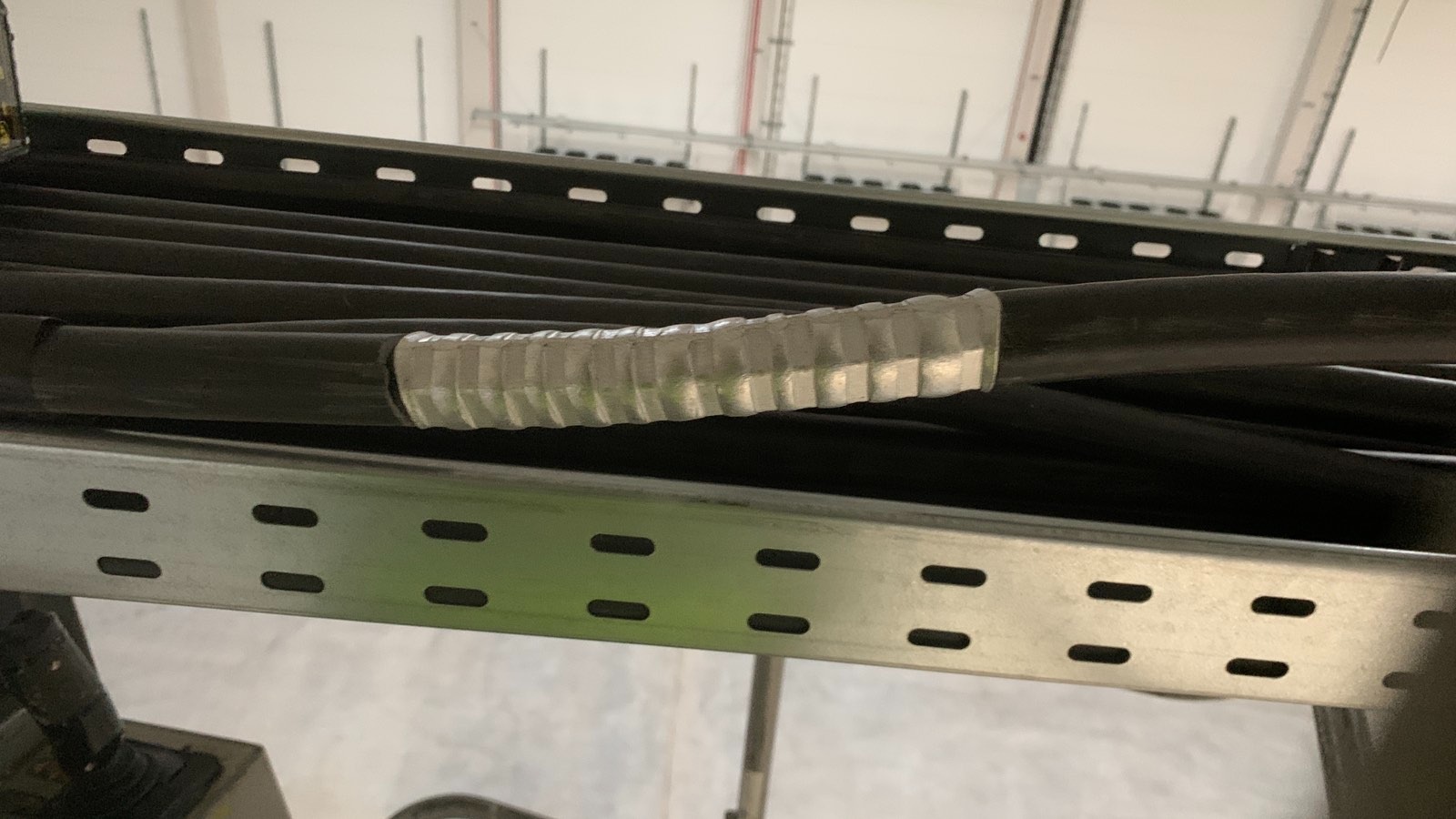 Rząd czarnych kabli elektrycznych, zabezpieczonych srebrną osłoną, ułożonych na metalowej perforowanej drabince kablowej, w jasnym, przemysłowym wnętrzu.