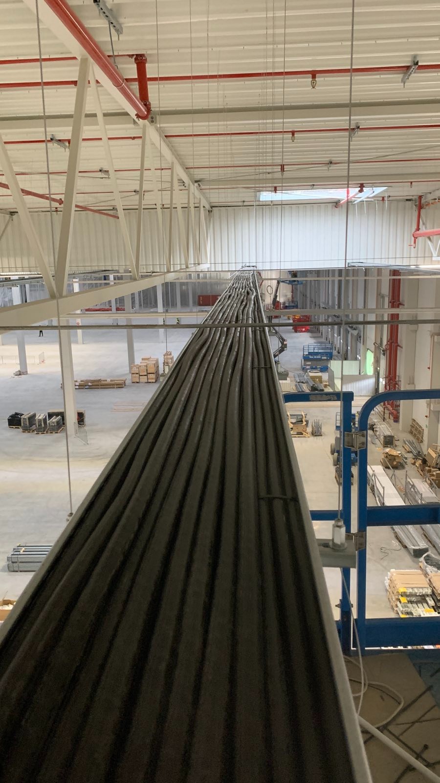 Wiązka kabli ułożona w korycie w hali przemysłowej, widok z góry. Widoczne elementy konstrukcji hali i podnośnik. Etap prac instalacyjnych.