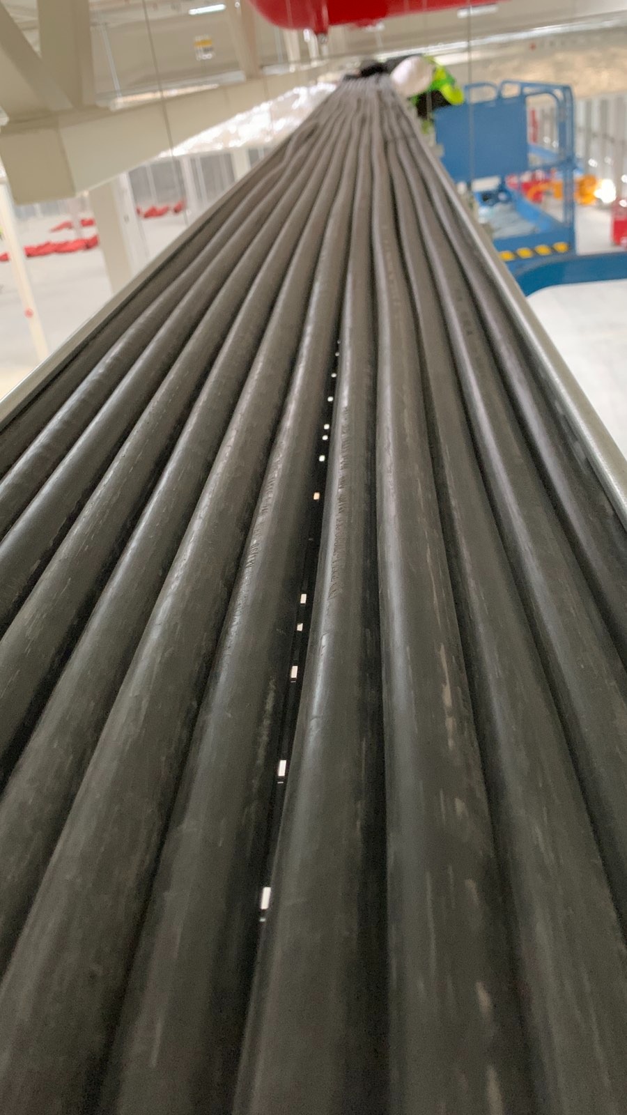 Równolegle ułożone, szare rury instalacji elektrycznej w hali przemysłowej, widoczne z dołu pod kątem. W tle podnośnik i elementy konstrukcji.