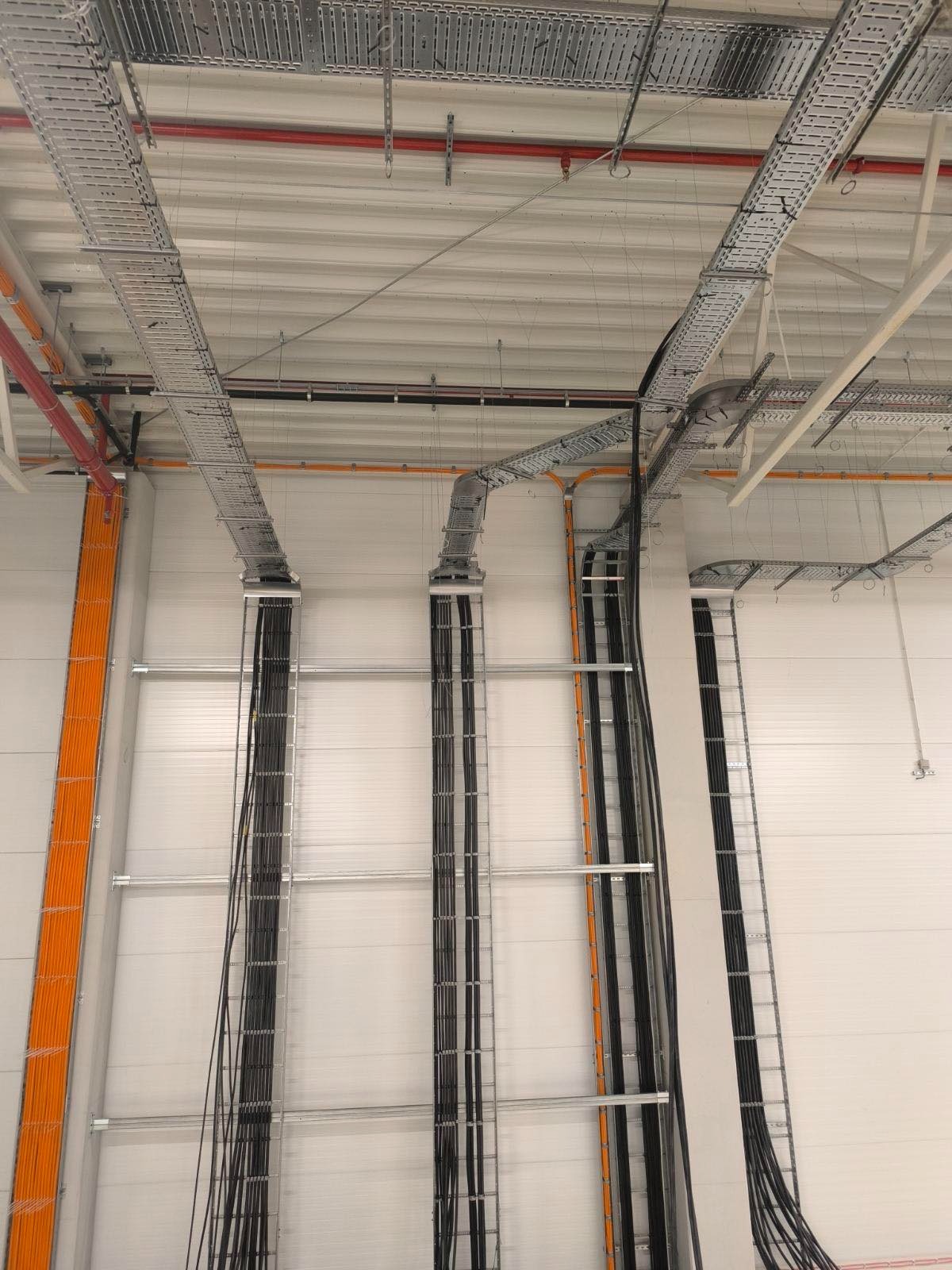 Wnętrze hali z widoczną, rozbudowaną instalacją kablową w metalowych korytach pod sufitem i wzdłuż ścian. Kable w kolorach czarnym i pomarańczowym.