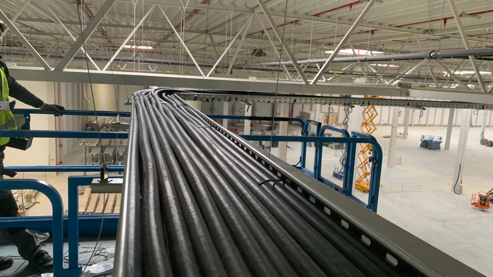 Rząd ułożonych kabli w korycie kablowym w hali produkcyjnej, widziane z podnośnika koszowego. W tle widoczna konstrukcja dachu i inne maszyny.