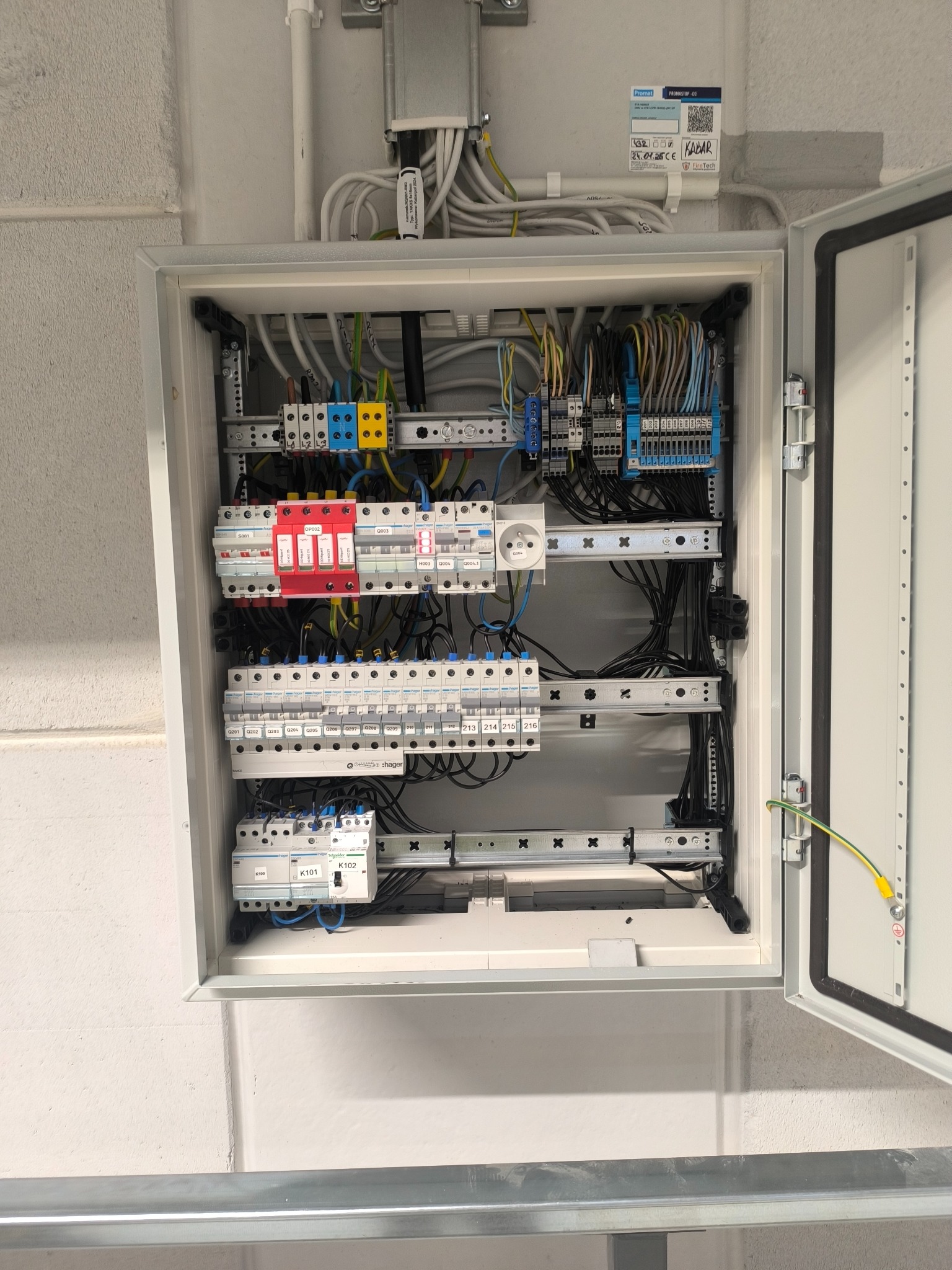 Otwarta skrzynka elektryczna z zainstalowanymi bezpiecznikami, przewodami i gniazdkiem na szarej ścianie. Widoczne detale instalacji, złącza i oznaczenia.