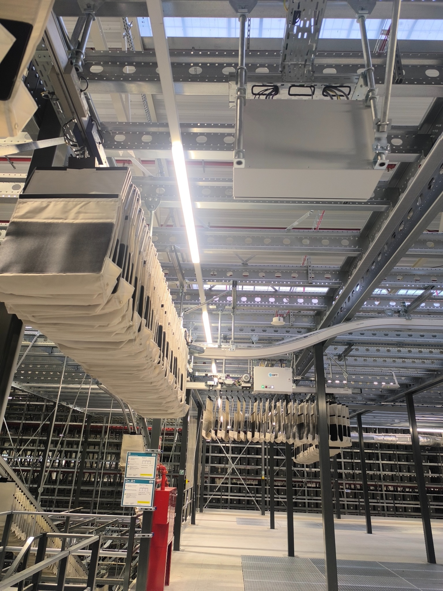 Oświetlenie LED w hali magazynowej z systemem transportu, widoczne kable i skrzynki elektryczne pod sufitem, metalowa konstrukcja nośna i regały magazynowe.