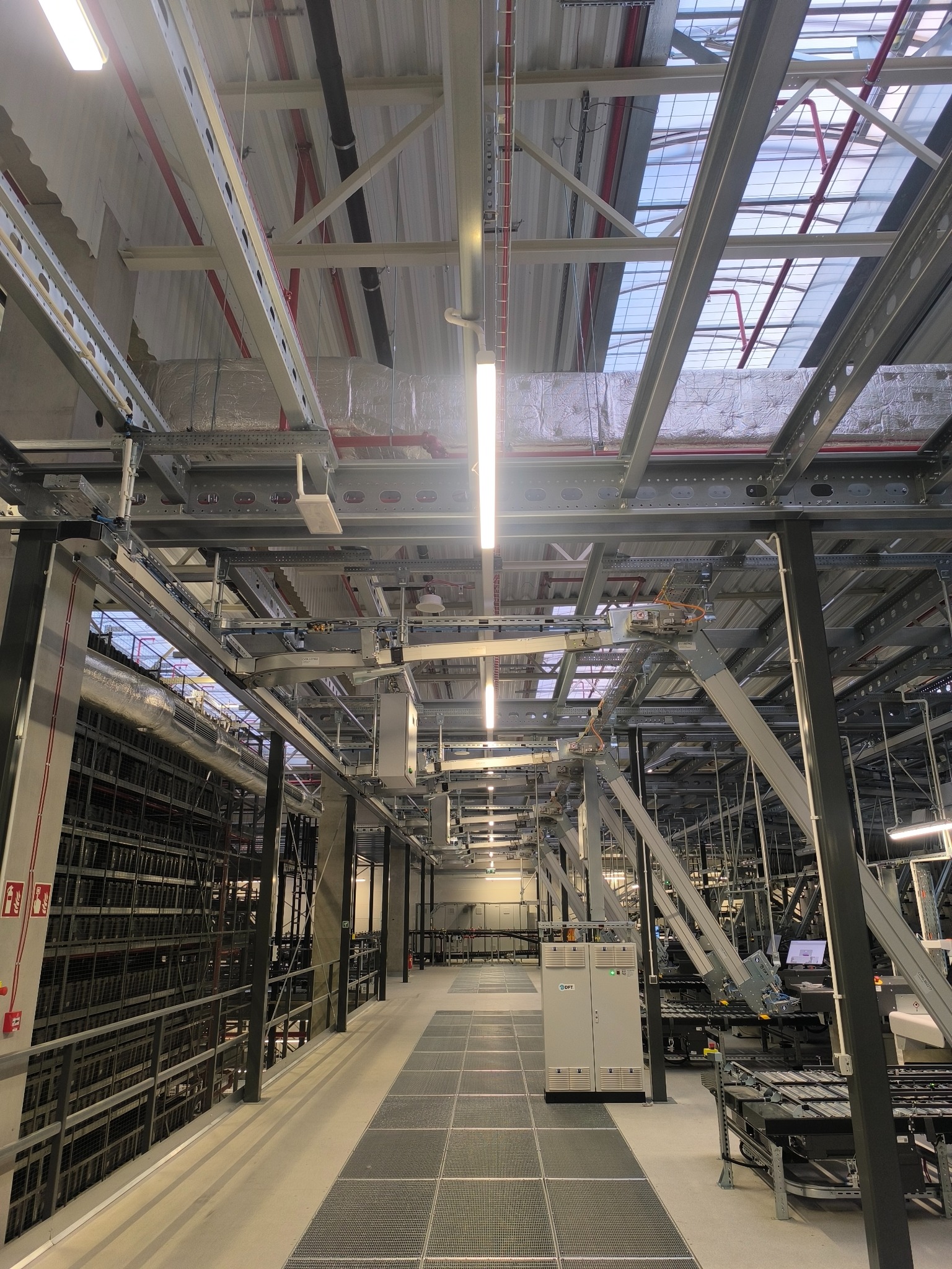 Wnętrze hali produkcyjnej z widocznymi instalacjami pod sufitem, oświetleniem liniowym i metalowymi konstrukcjami. Perspektywa z dołu ukazuje złożoność infrastruktury.