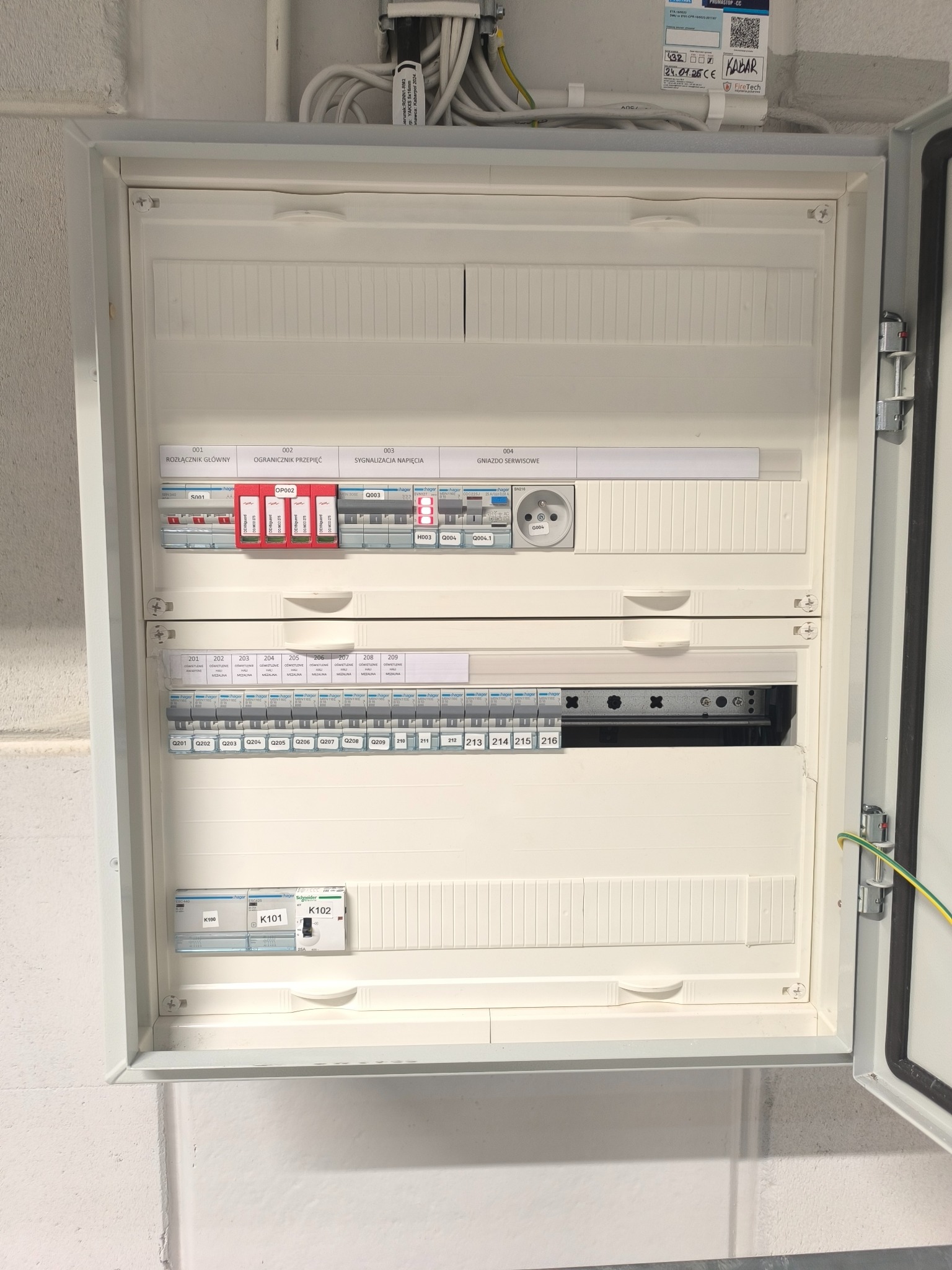 Otwarta, biała skrzynka elektryczna z bezpiecznikami, wyłącznikami i gniazdem serwisowym. Widoczne oznaczenia na elementach, przewody i etykieta z datą.