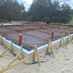 Zbrojone fundamenty budynku w trakcie budowy, widoczne szalunki, izolacja termiczna, siatka zbrojeniowa i pionowe rury instalacyjne. W tle drzewa i osoba.