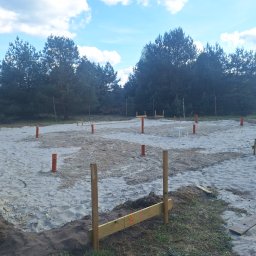 Firma budowlana Jacek Barszczewski - Przygotowany teren pod fundamenty z widocznymi deskami szalunkowymi i pomarańczowymi rurami wentylacyjnymi na piaszczystym podłożu, w tle las i błękitne niebo.