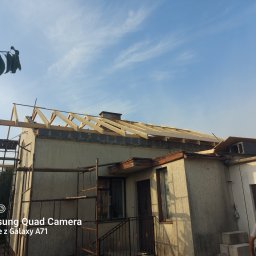 Firma budowlana Jacek Barszczewski - Montaż więźby dachowej na jednopiętrowym domu z pomocą rusztowania i drabiny, widok z dołu na błękitne niebo. Prace dekarskie w toku.