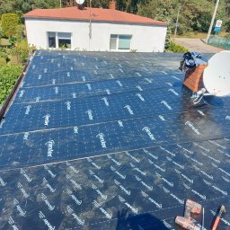 Firma budowlana Jacek Barszczewski - Dach płaski pokryty czarną membraną hydroizolacyjną z widocznym kominem, anteną satelitarną i narzędziami dekarskimi. Widok na okoliczne budynki i zieleń.