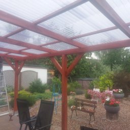 Firma budowlana Jacek Barszczewski - Drewniana pergola z przezroczystym zadaszeniem w ogrodzie, drabina oparta o konstrukcję, narzędzia, roślinność w tle, widok z dołu na zadaszenie.