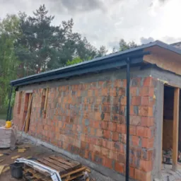 Budowa domu w trakcie: murowane ściany z pustaków ceramicznych, zamontowane rynny, widoczna więźba dachowa. Elementy konstrukcyjne i materiały budowlane na placu budowy.