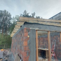 Firma budowlana Jacek Barszczewski - Budowa niskiego budynku z czerwonej cegły, widoczne drewniane elementy konstrukcyjne dachu i narzędzia na stole wewnątrz. W tle las i fragment budynku z szarego kamienia.