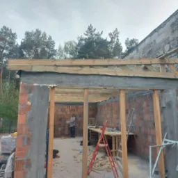 Budowa garażu z czerwonej cegły: wzniesione mury, drewniana konstrukcja dachu w trakcie montażu, drabiny i narzędzia budowlane wewnątrz. Praca w toku.