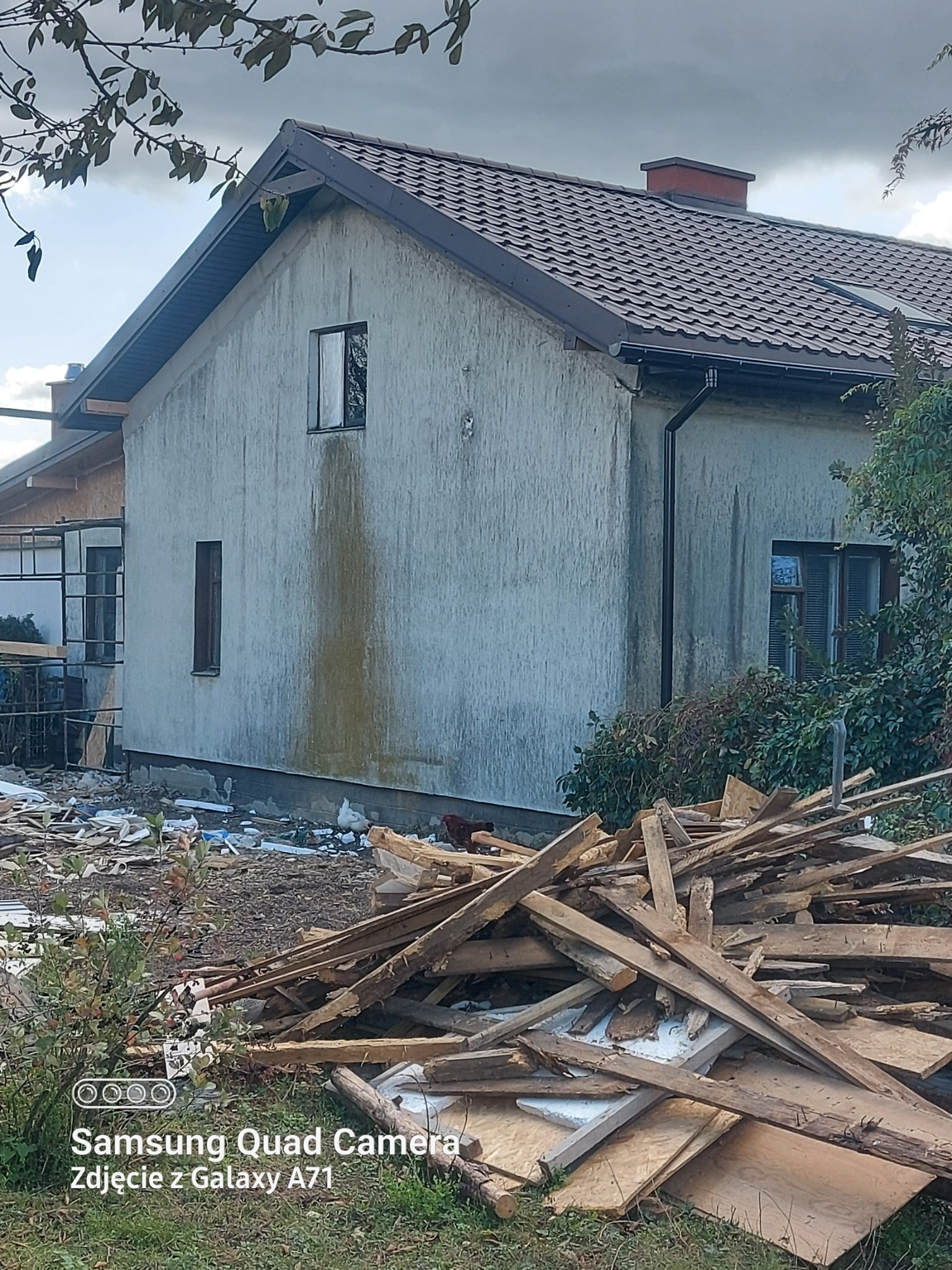 Dom z szarą elewacją i brązowym dachem z blachodachówki. Sterta drewna i gruzu przed budynkiem. Widoczne ślady zacieków na ścianie.