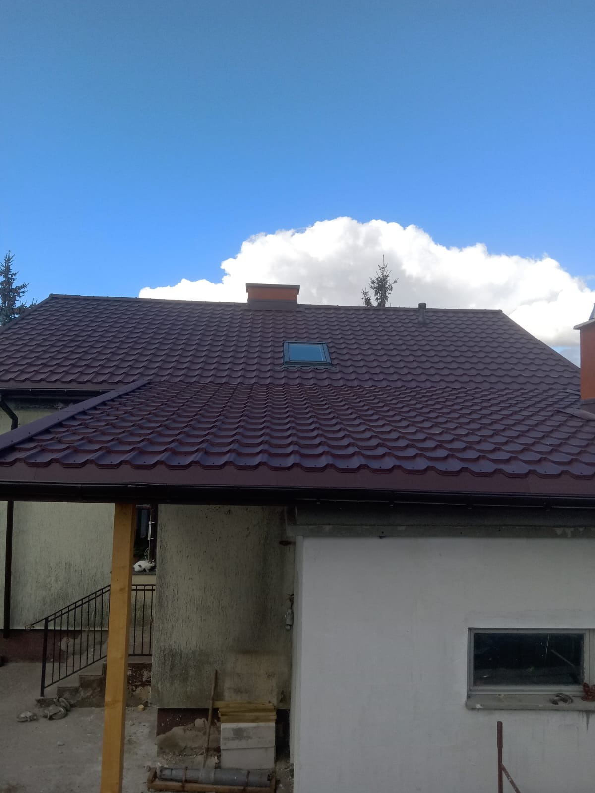 Ciemnoczerwony dach z blachodachówki z oknem dachowym, kominem i rynną, na tle błękitnego nieba z białymi obłokami. Widoczny fragment elewacji budynku.