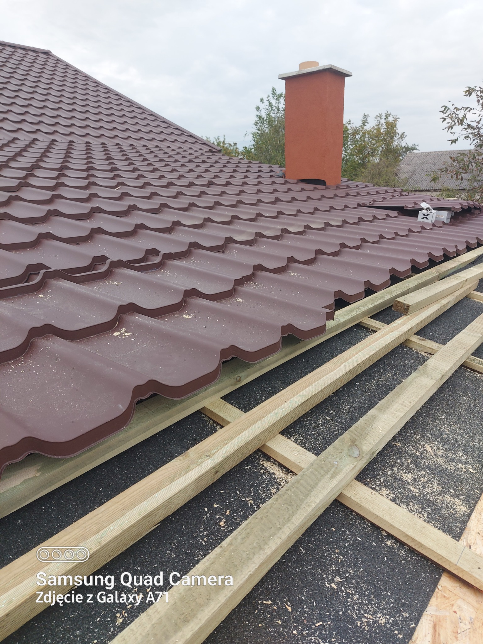 Częściowy widok brązowego dachu z blachodachówki trapezowej w trakcie montażu. Widoczna więźba dachowa i komin. Fragment dachu pokryty, reszta w trakcie prac.