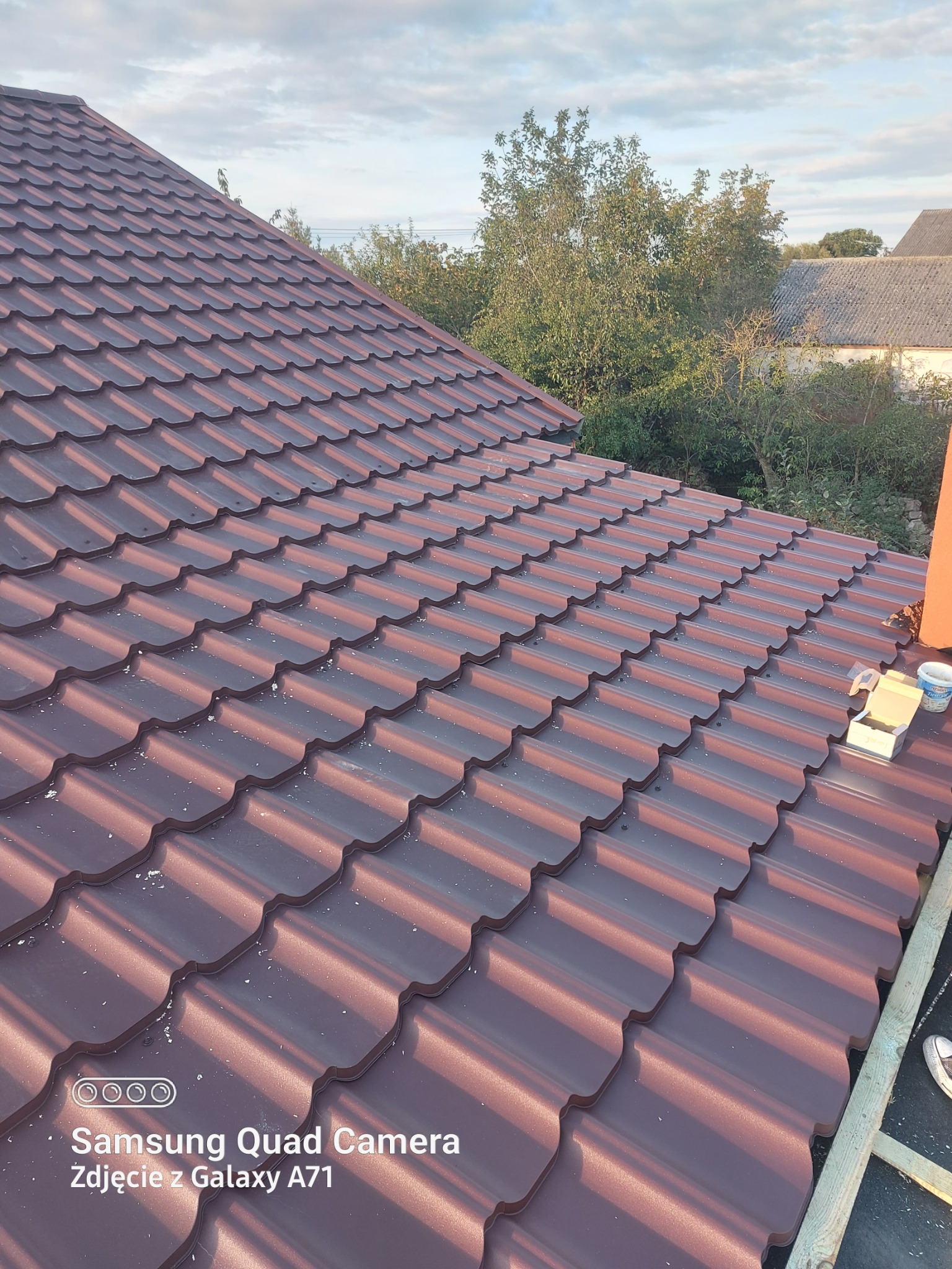 Ciemnobrązowy dach z blachodachówki modułowej, widok z góry, z elementami montażowymi i fragmentem buta w prawym dolnym rogu. W tle zieleń drzew i fragment dachu sąsiedniego budynku.