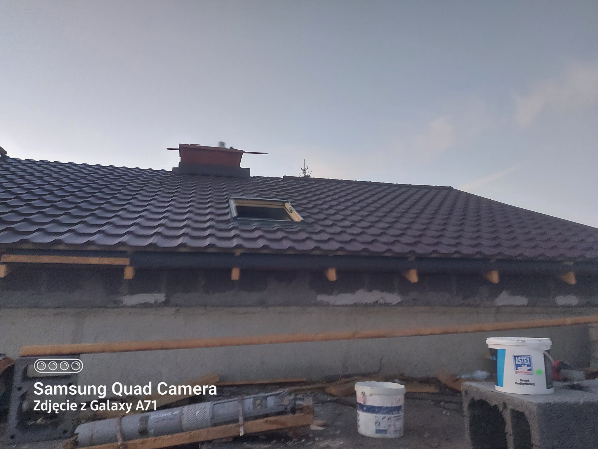 Brązowy dach z dachówką trapezową i oknem dachowym w trakcie montażu, widoczny komin, ściany budynku w stanie surowym, farba gruntująca Astex w wiadrze.