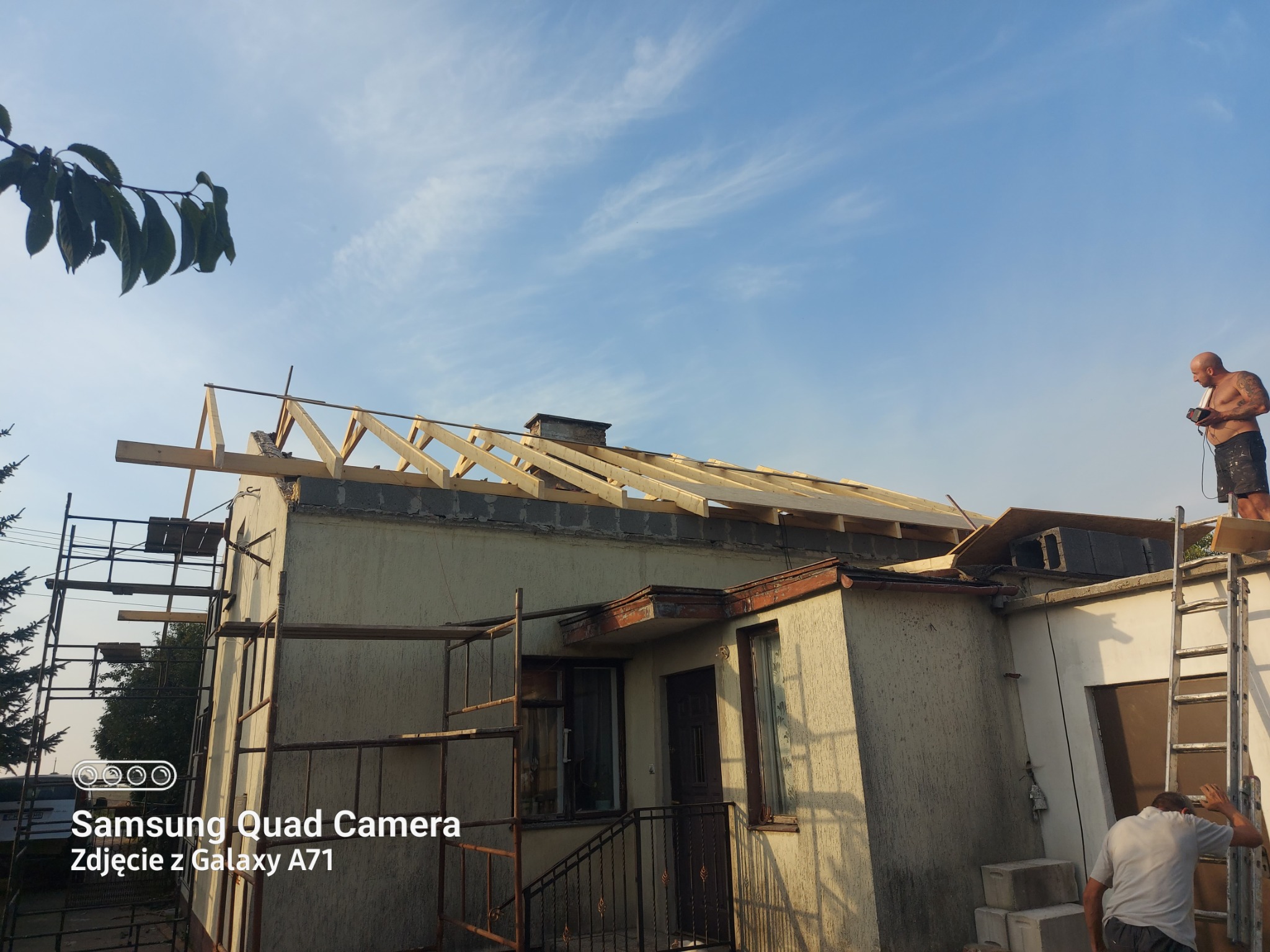 Montaż więźby dachowej na jednopiętrowym domu z pomocą rusztowania i drabiny, widok z dołu na błękitne niebo. Prace dekarskie w toku.