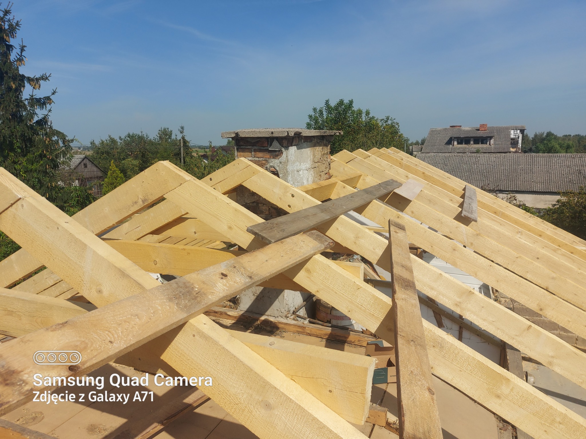 Więźba dachowa z jasnego drewna w trakcie montażu, widoczny komin i fragmenty okolicznych budynków w tle, słoneczny dzień.