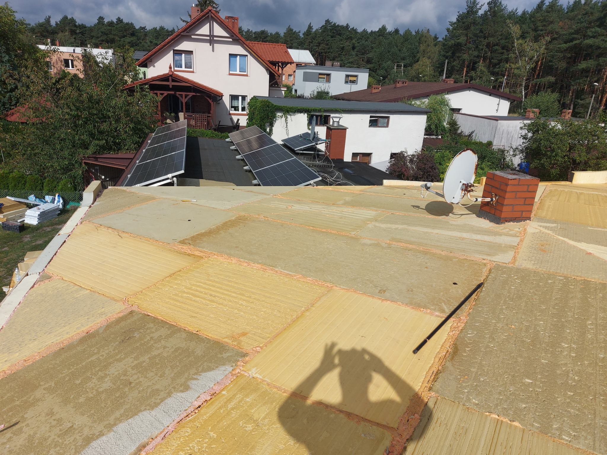 Ocieplenie dachu pianą PUR w trakcie prac. Widoczne panele słoneczne na sąsiednim dachu oraz cień osoby wykonującej zdjęcie. Dach z widokiem na zabudowę jednorodzinną.