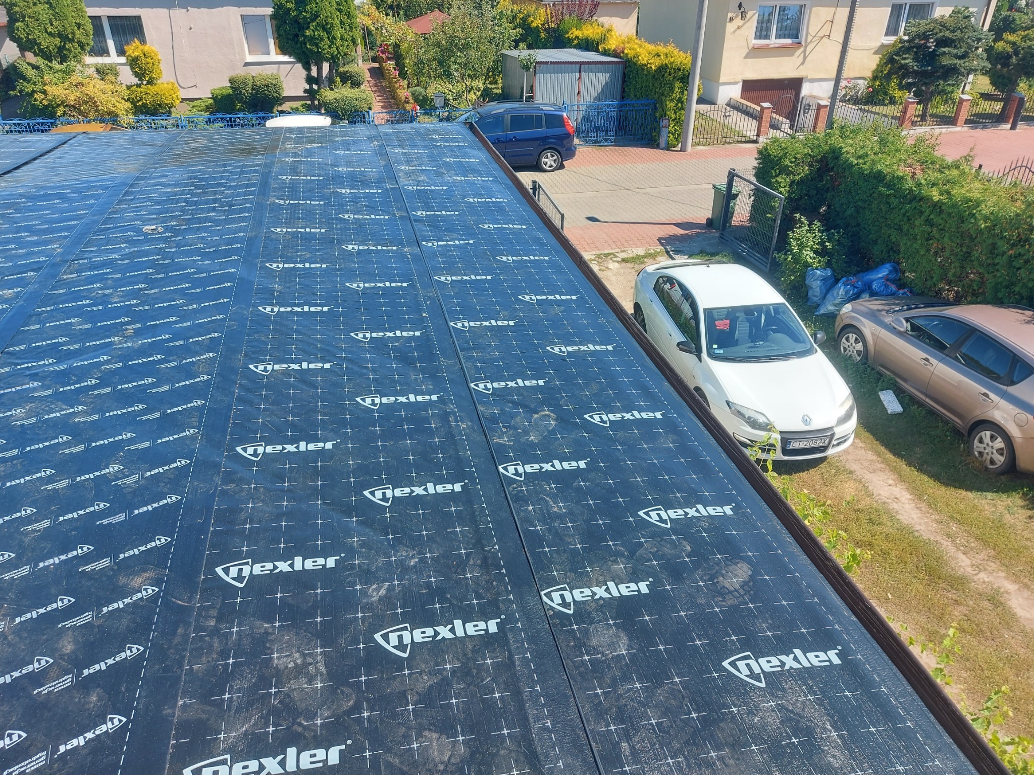 Widok z góry na dach pokryty czarną membraną hydroizolacyjną z nadrukiem, w tle domy i samochody, wykonane obróbki blacharskie.