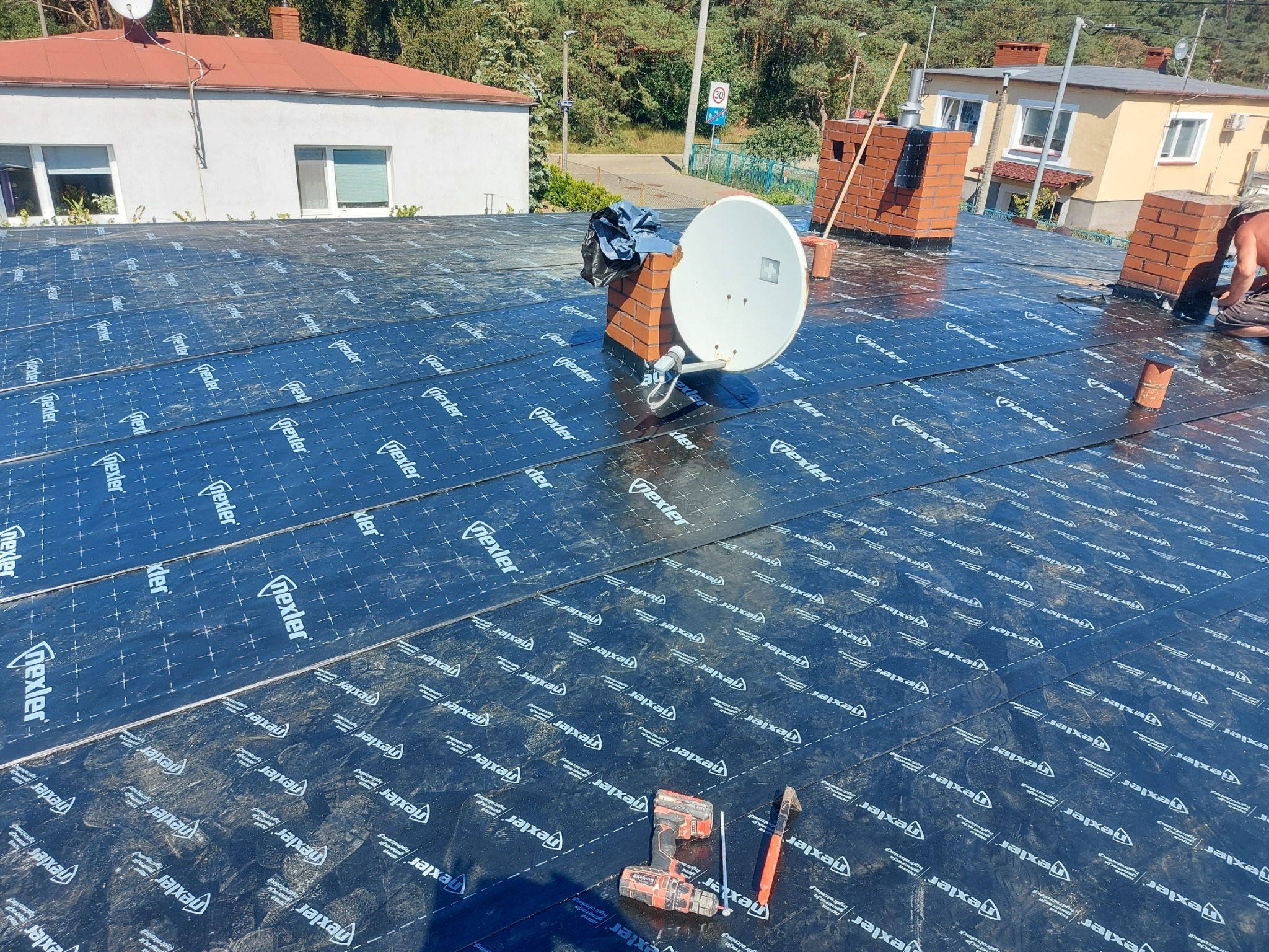 Dach pokryty membraną hydroizolacyjną Nexler, widoczne narzędzia (wkrętarka, nóż), antena satelitarna i kominy. Prace dekarskie w toku, widok z góry.