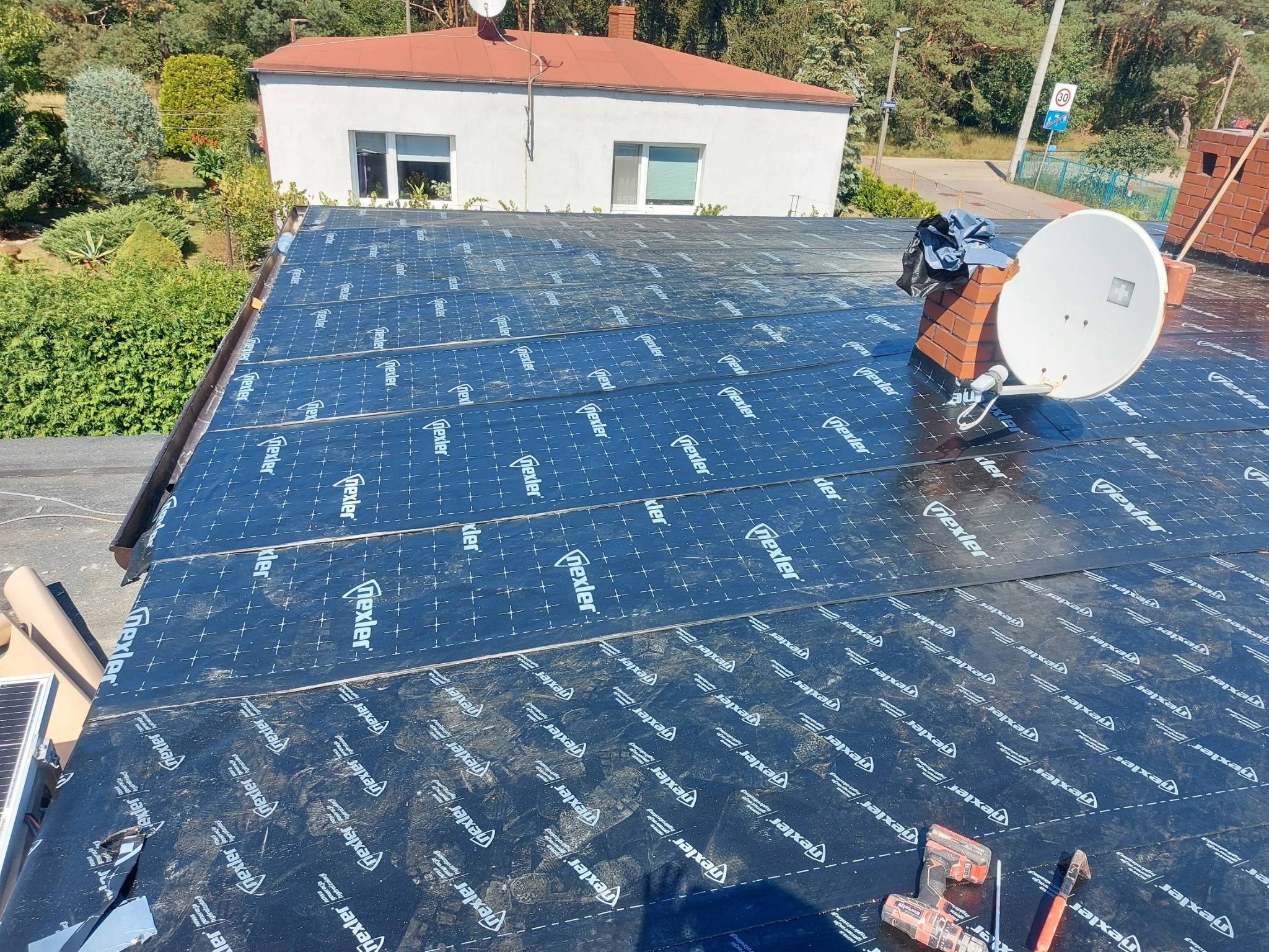 Dach płaski pokryty czarną membraną hydroizolacyjną z widocznym kominem, anteną satelitarną i narzędziami dekarskimi. Widok na okoliczne budynki i zieleń.