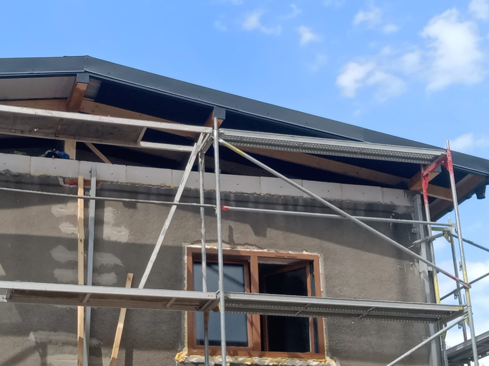 Dom w trakcie budowy: widoczne rusztowania, surowy tynk, drewniana rama okienna i fragment dachu z czarnym orynnowaniem na tle błękitnego nieba.