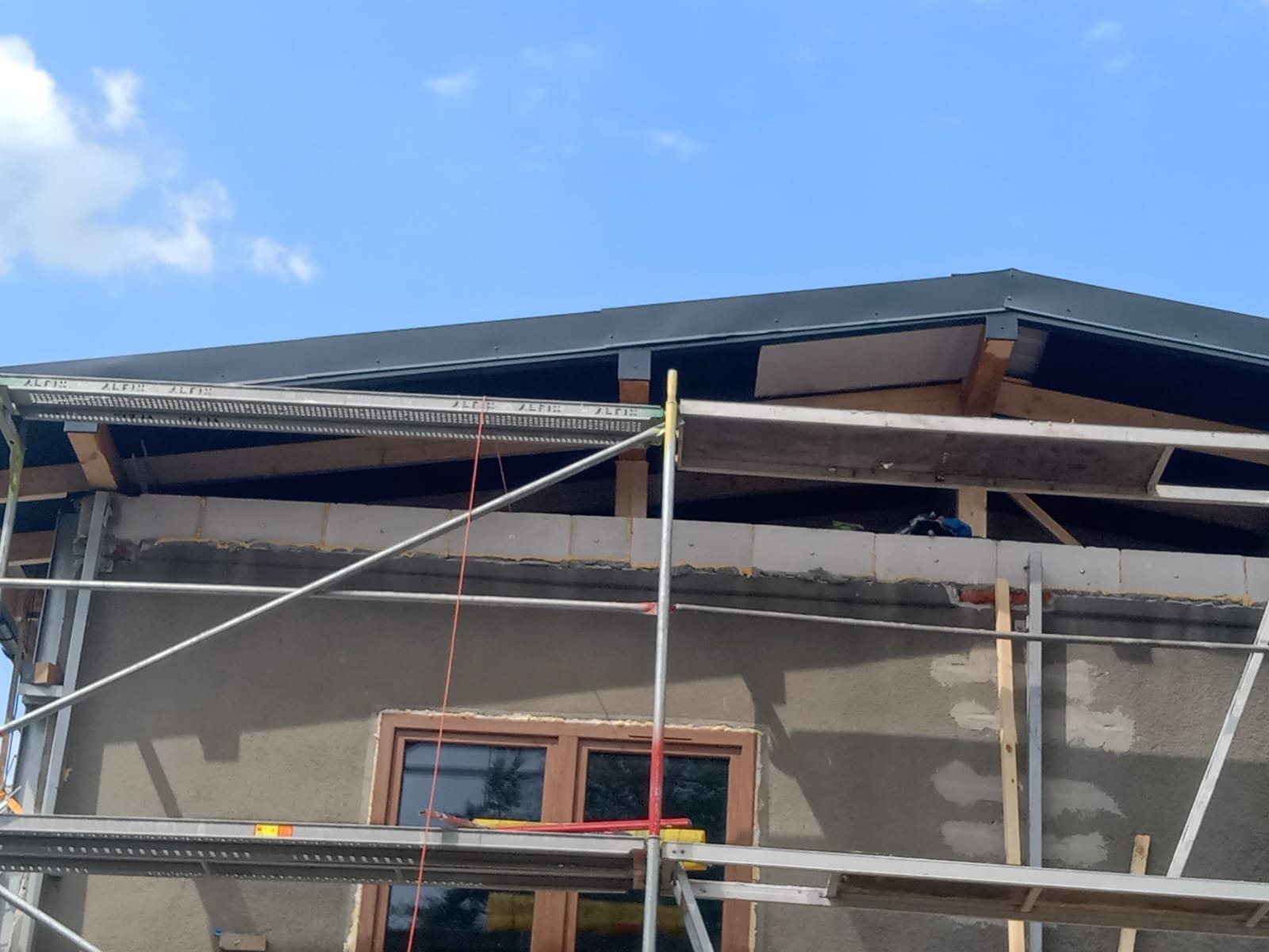 Dach z czarną blachodachówką w trakcie montażu, widoczny ze rusztowania. Budynek w trakcie prac wykończeniowych elewacji. Błękitne niebo w tle.